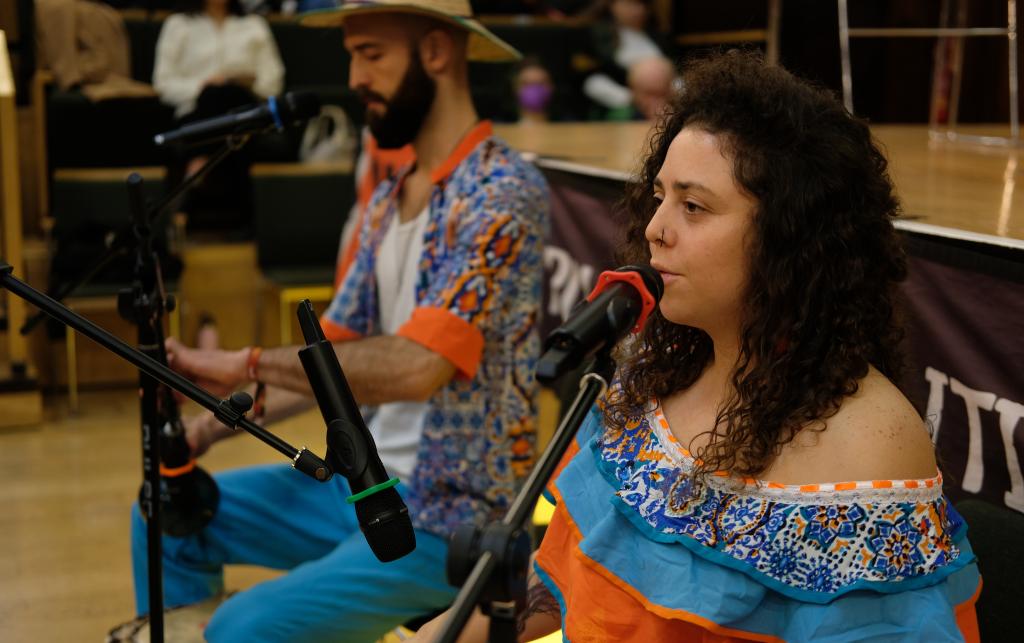 A close up of a man and a woman in the middle of a performance. Both have microphones in front of them which they appear to be singing into. The man is also holding a guitar. 