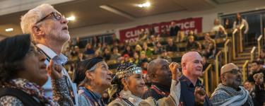 Photo of speakers standing in a row, looking upwards. Some have their fists raised in the air. Behind them we can see crowds of people sitting in an auditorium.