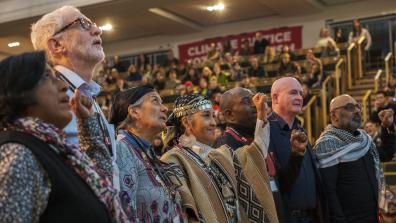 Photo of speakers standing in a row, looking upwards. Some have their fists raised in the air. Behind them we can see crowds of people sitting in an auditorium.